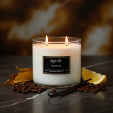 Load image into Gallery viewer, A three-wick scented candle labeled BOUGIE NAPA 'Pick Me Up' sits on a dark marble surface with coffee beans, vanilla beans, a lemon wedge, and dried leaves around it. The candle is lit with two visible flames against a warm blurred background.