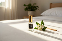 Load image into Gallery viewer, Bougie Napa Fresh room and linen spray bottle with gold cap sitting on a sunlit white bed next to fresh white citrus blossoms.
