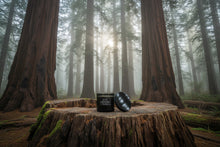 Load image into Gallery viewer, Redwood Grove candle in dark green tinted glass sitting in a misty redwood grove.
